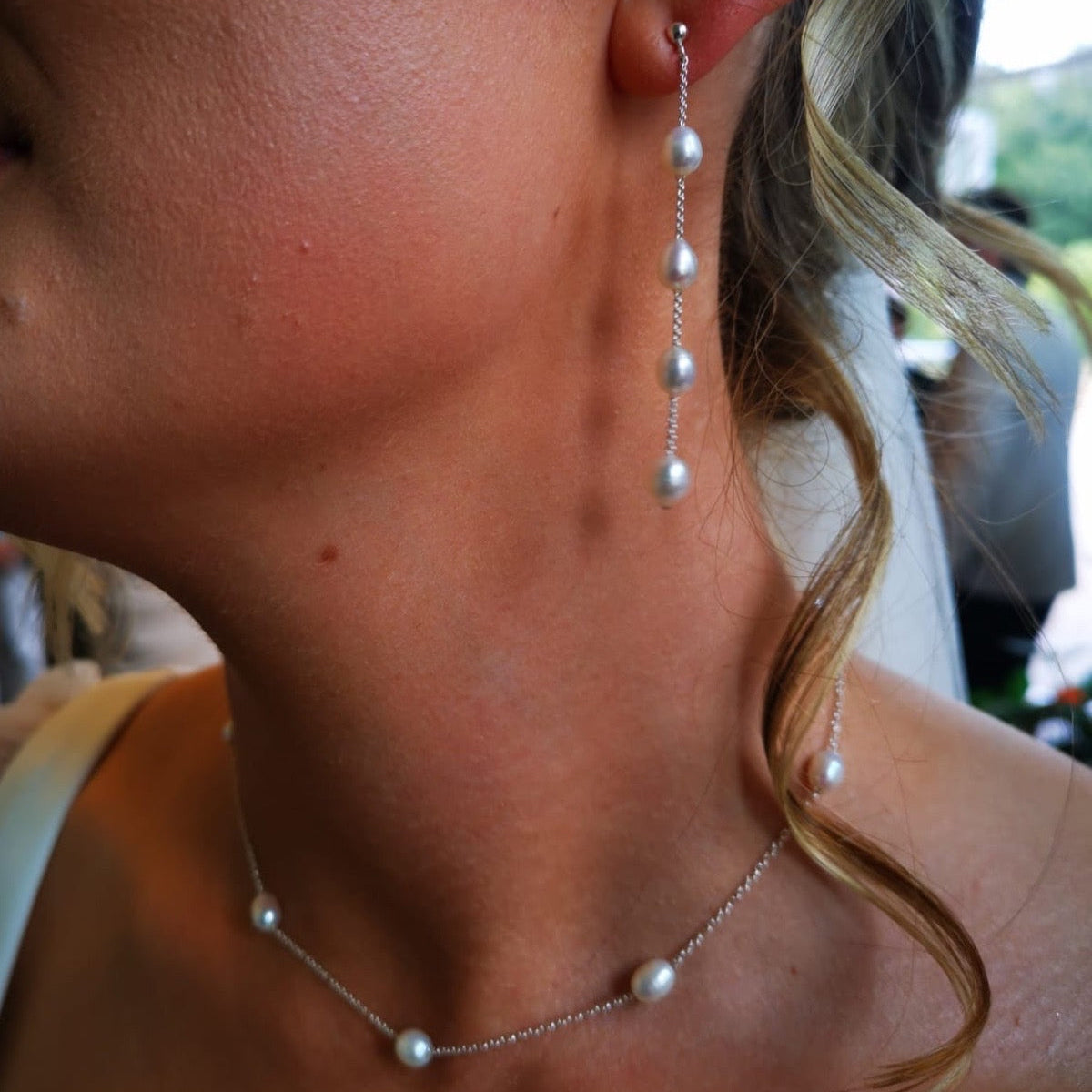 A close-up lifestyle shot of a woman wearing a delicate silver chain necklace with white pearls and a matching long drop earring with three dangling pearls.